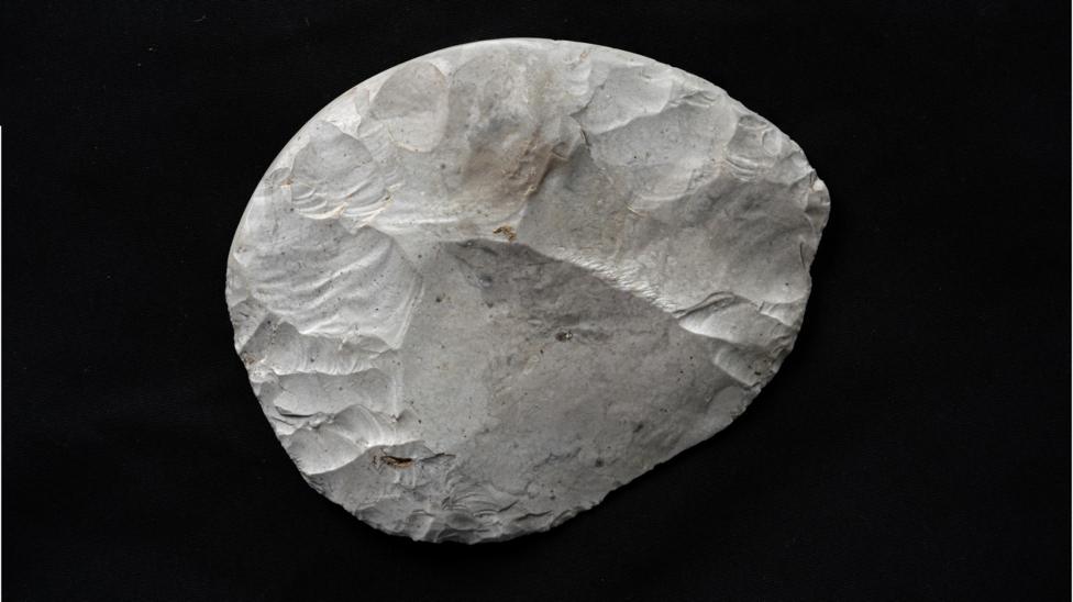 A rounded piece of flint that has been carefully worked to create a knife. It is white, with some brown and grey marks, and is set against a black background. The areas where pieces of the flint have been struck off using another stone are clearly visible. It is not a perfect circle but its sides are rounded. 