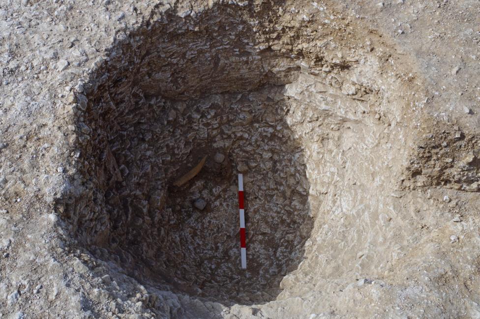 The hole in the ground shows a light rock with a hole in shadow dug into it. At the bottom is a red and white measuring pole, with three blocks of white and two blocks of red visible in between. It's difficult to see how deep it is but at the bottom there is an artefact protruding at the bottom on the left hand side of the hole. It is in the shadowed area and is something that looks light brown in colour and slightly curved. 
