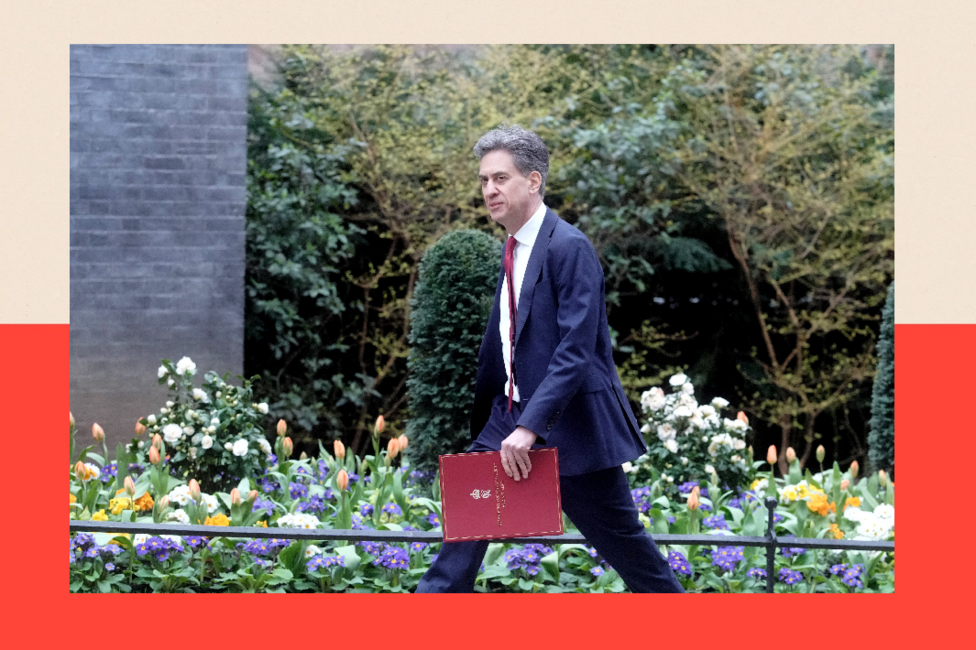 Ed Miliband walking in Downing Street, carrying a red briefcase. There are slpring flowers in bloom behind him, including orange tulips.