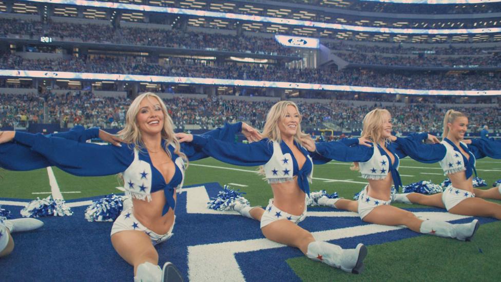 The Dallas Cowboys Cheerleaders in formation on the football field with crowds behind them. Four blonde cheerleaders look straight ahead with their legs flat, performing the splits.