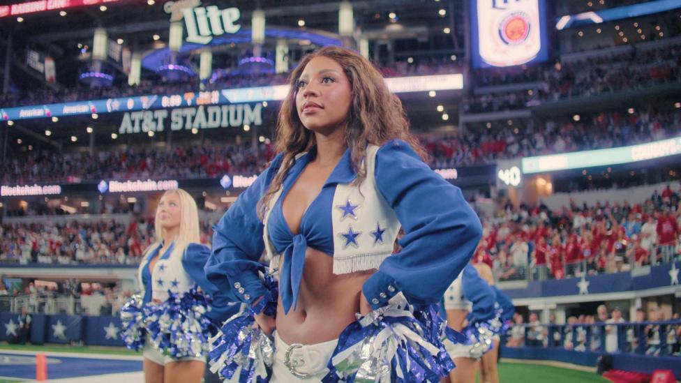 Two dancers on the field look straight ahead. The dancer in the front has long brown hair and the dancer in the back has long straight blonde hair. They wear the signature blue and white Dallas Cowboys Cheerleaders uniform