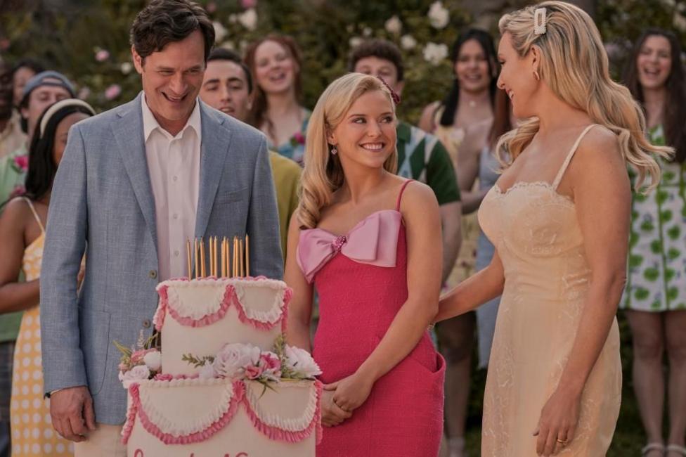 A man and two women smiling at each other, standing infront of a two-tierd cake, with gold candles and pink decorations. People are in the background, also smiling.
