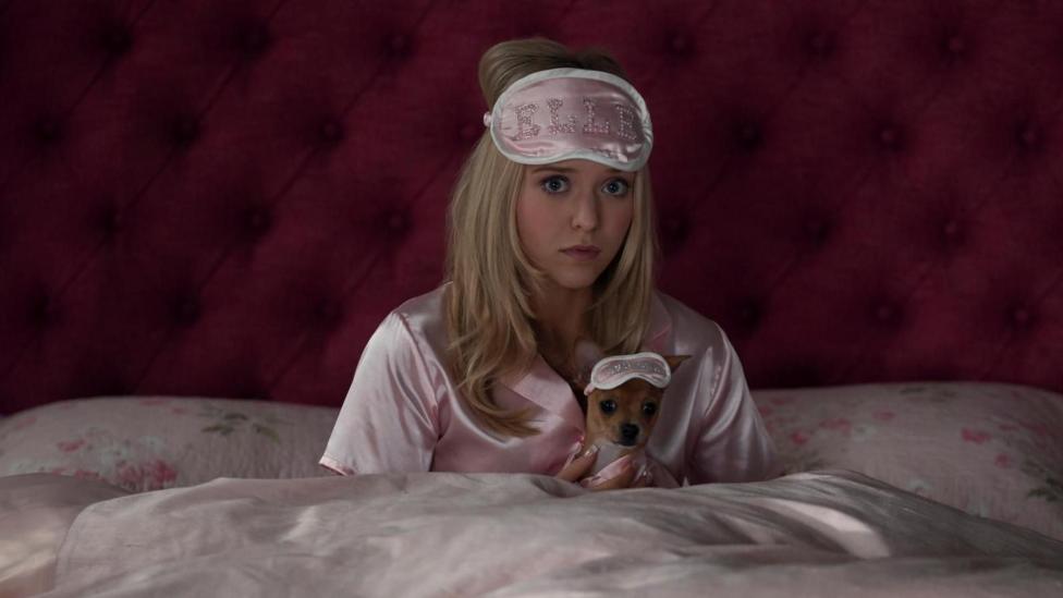 A woman with long blonde hair sits up in bed, with pink bedsheets, pink satin PJs and a pink eye mask on her forehead that has