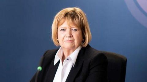 Baroness Hallett in front of a blue backdrop featuring the pink, purple and white Covid-19 inquiry logo.