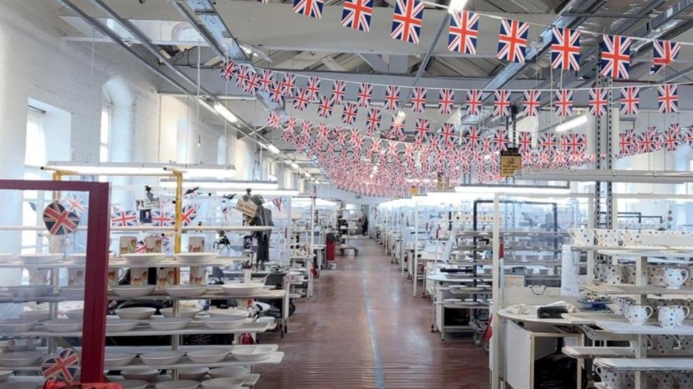 Inside a warehouse. The walls are painted white with big windows letting in the light. Strings of small Union Jack flags hang from the ceiling. A corridor goes down the middle of the room and on each side are rows of mugs and plates.
