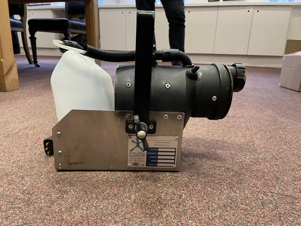 A mechanical snow machine sitting on a carpet. It comprises of a stainless steel box with black and plastic components.