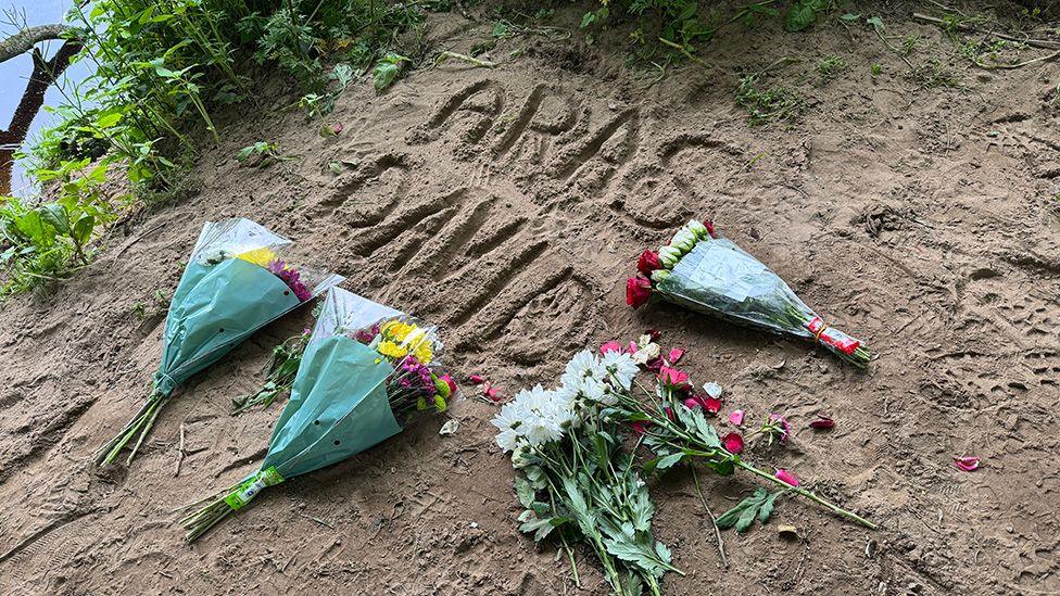 Bouquets of flowers on the ground of the river bank. The names 'Aras' and 'David' are drawn in the sand.