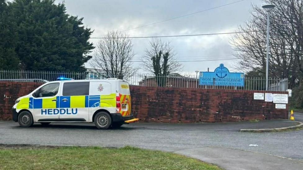A brown school wall with a silver railing above and a police van parked outside. In the background is a blue school sign.