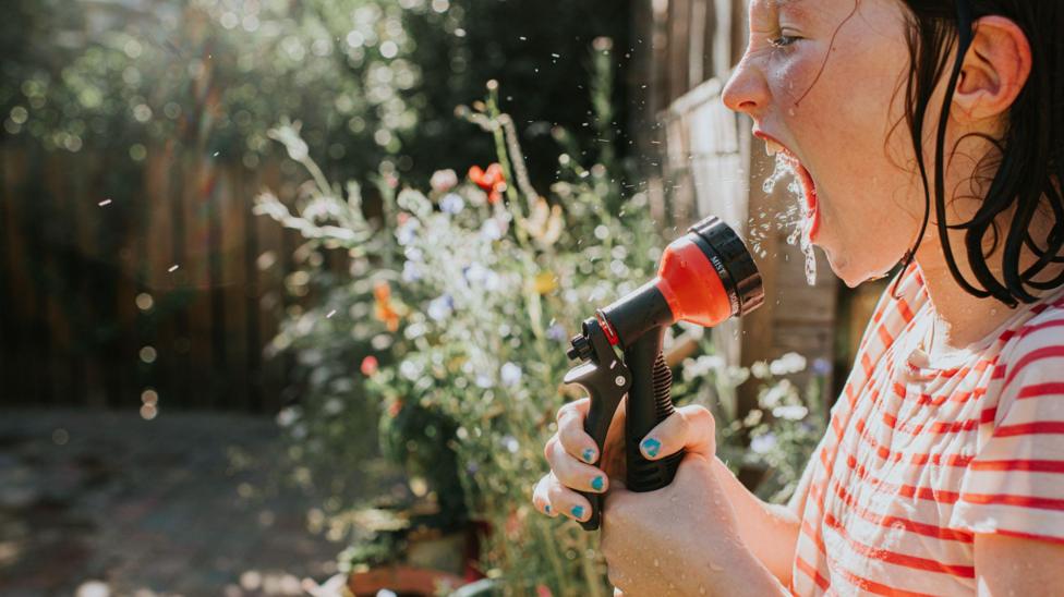 A little girl sprays water directly into her mouth using a hosepipe - stock photo A child attempts to cool off on a very hot day using a hose pipe. She sprays water directly into her mouth.