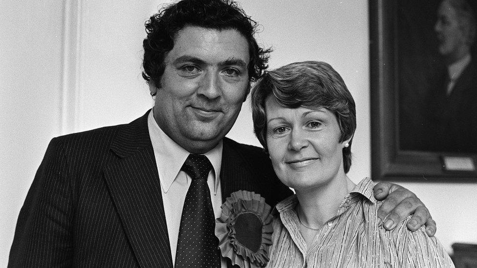 John and Pat Hume are pictured together. He is on the right, wearing a suit and large rosette. Pat, on the left is smiling. His arm is on her shoulder.