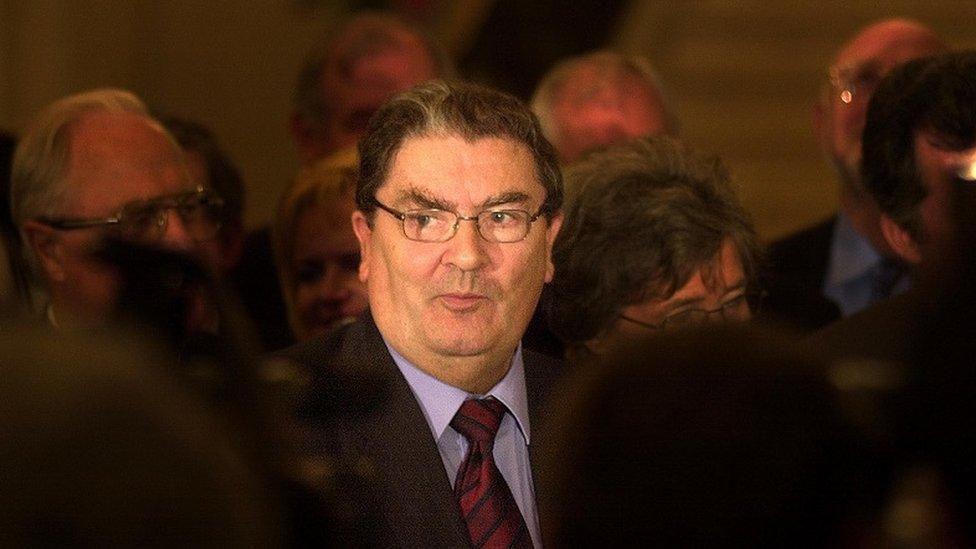 John Hume stands among a crowd of people. He is wearing glasses and has short dark hair. He is wearing a suit, shirt and tie