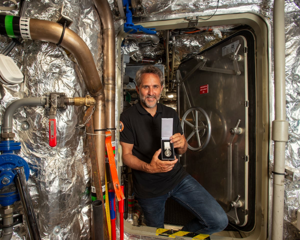 A bearded older man poses with a medal in a bulkhead door of a chamber lined with padded silver foil and pipes