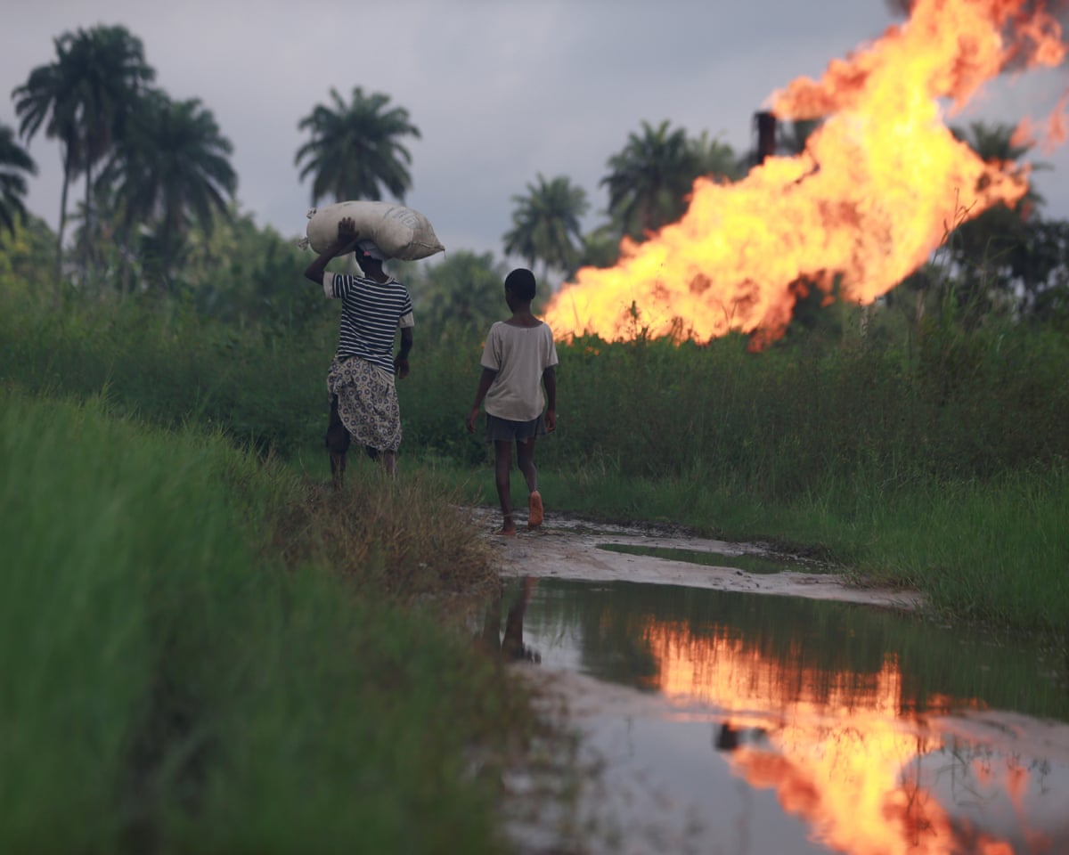 A boy follows a woman carrying a sack on her head as they walk along a riverbank in a tropical setting towards a burst of flame