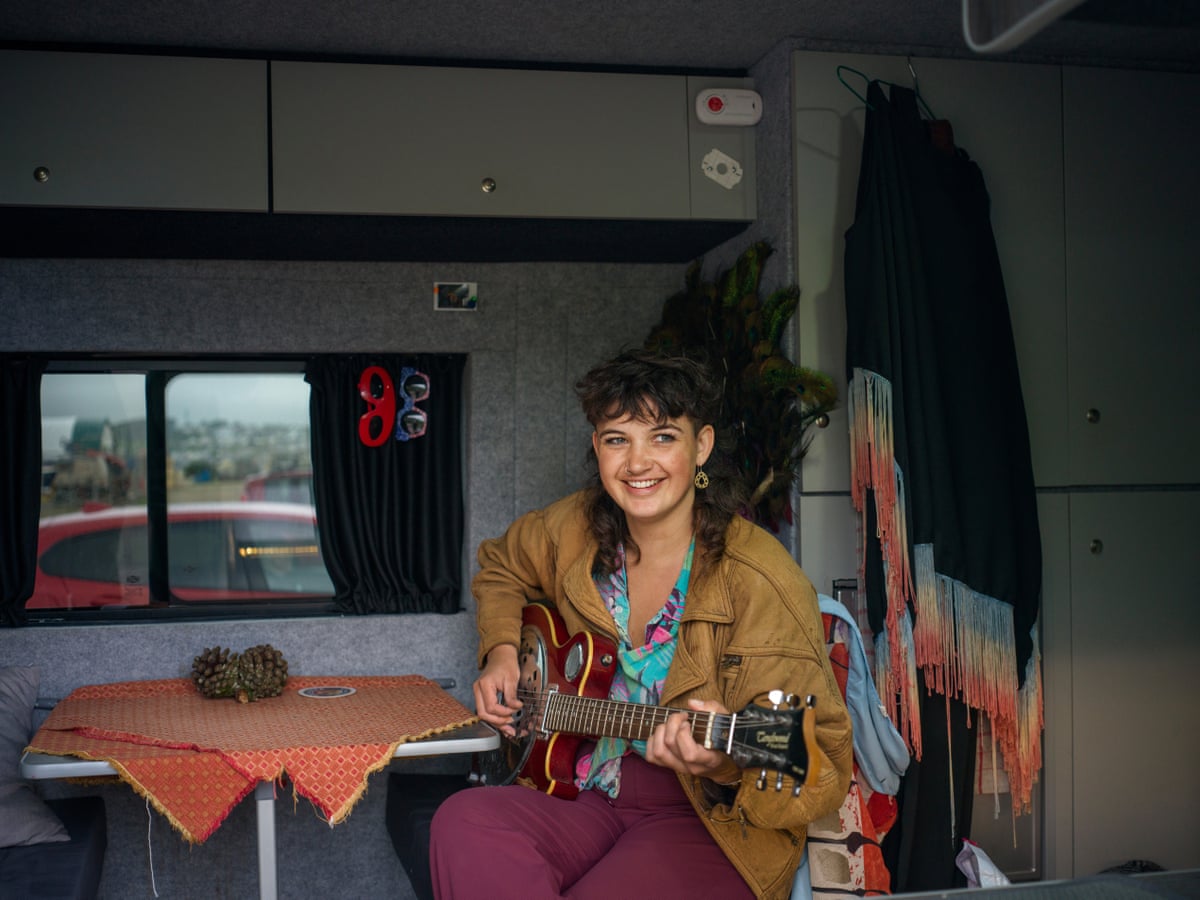 A young woman sits at a small table inside a van, holding a guitar.