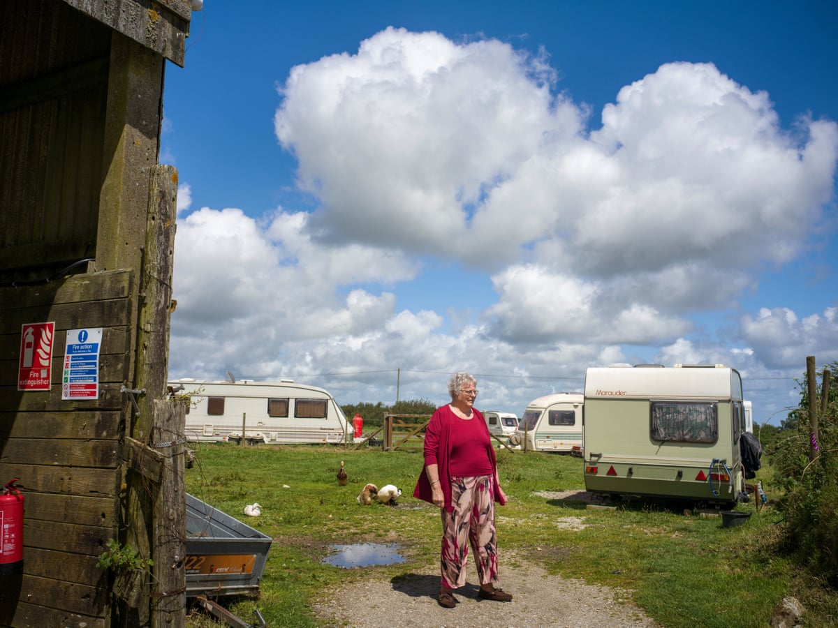 A woman stands in a field in which poultry roam and a few old caravans are parked.