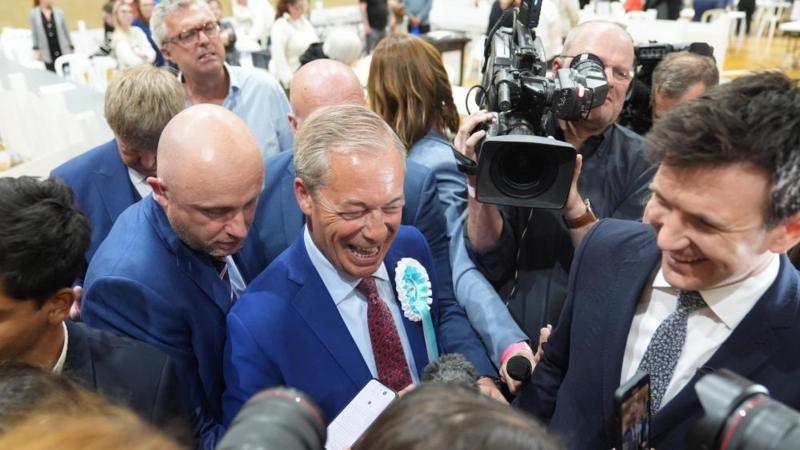 A tightly-packed crowd of men, some holding cameras. The none at the centre has grey hair. He is wearing a blue suit and shirt with a red tie and blue and white rosette and is smiliing.