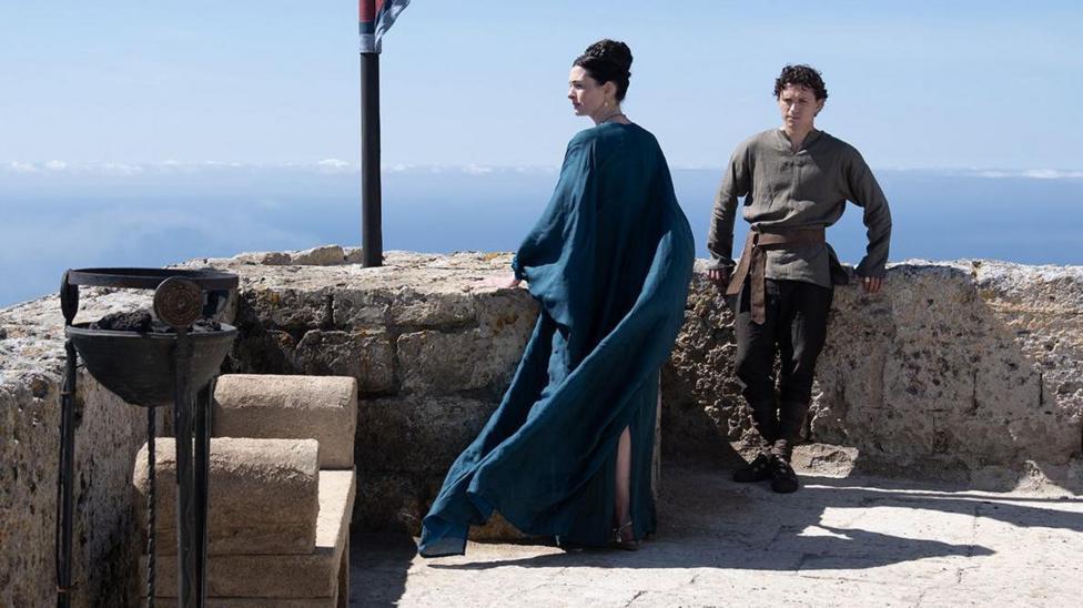 Anne Hathaway is wearing a flowing blue dress and Tom Holland a brown shirt and dark leggings. The actors are standing in sunshine at the battlements of a castle.