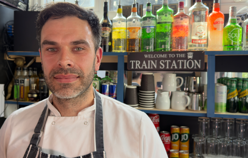 A white man with dark hair and stubble wearing a white food preparation coat and a blue and white stripey pinny in front of shelves full of drink