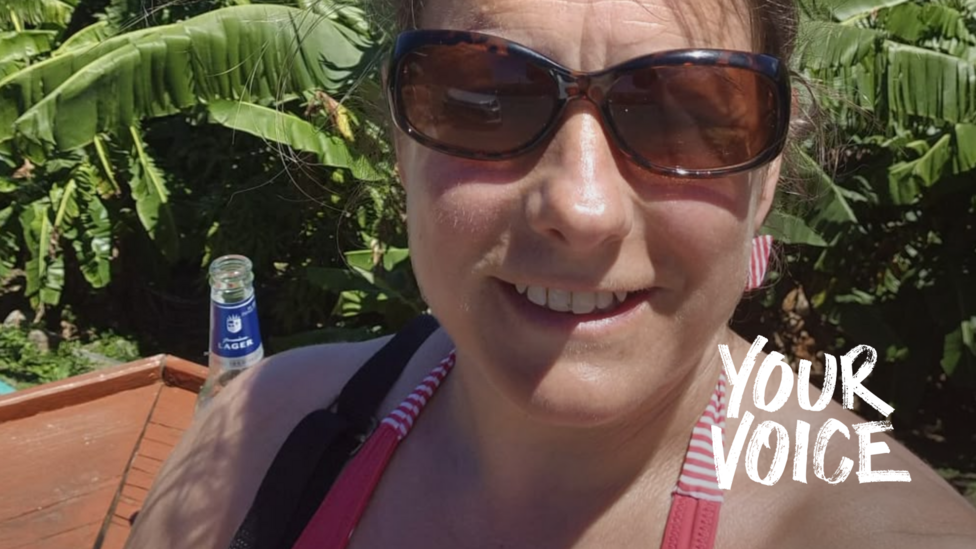 Flora Badger, wearing a red halter top and dark brown sunglasses, smiles taking a selfie. Behind her there is a view of the sea.
