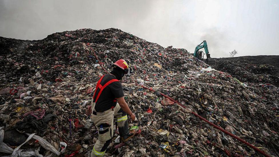 A person in red suspenders and a red helmet walks up a gigantic pile of rubbish. An excavator machine is in the background