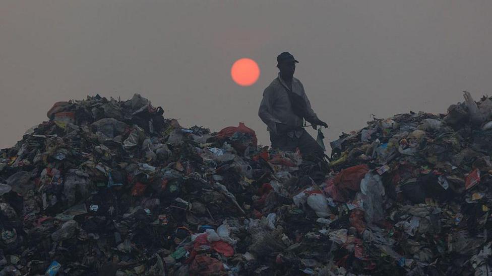 A person walks amid piles of rubbish with a low red sun in the sky behind him