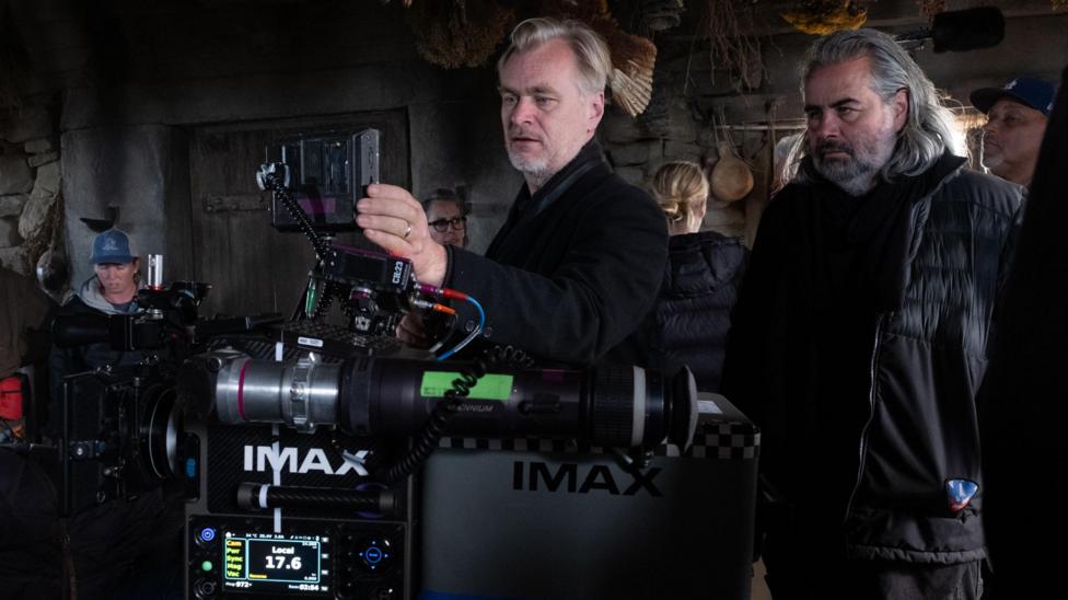 Christopher Nolan in the centre of the photo with cinematographer Hoyte van Hoytemaon on the right. Nolan is looking through Imax camera. Both are wearing dark clothes.