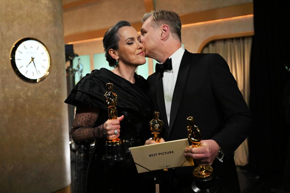 On the right -- Christopher Nolan holding two Oscars and Best Picture envelope, in black dinner jacket kissing his wife on the cheek On the left - Emma Thomas in black dress holding Oscar 