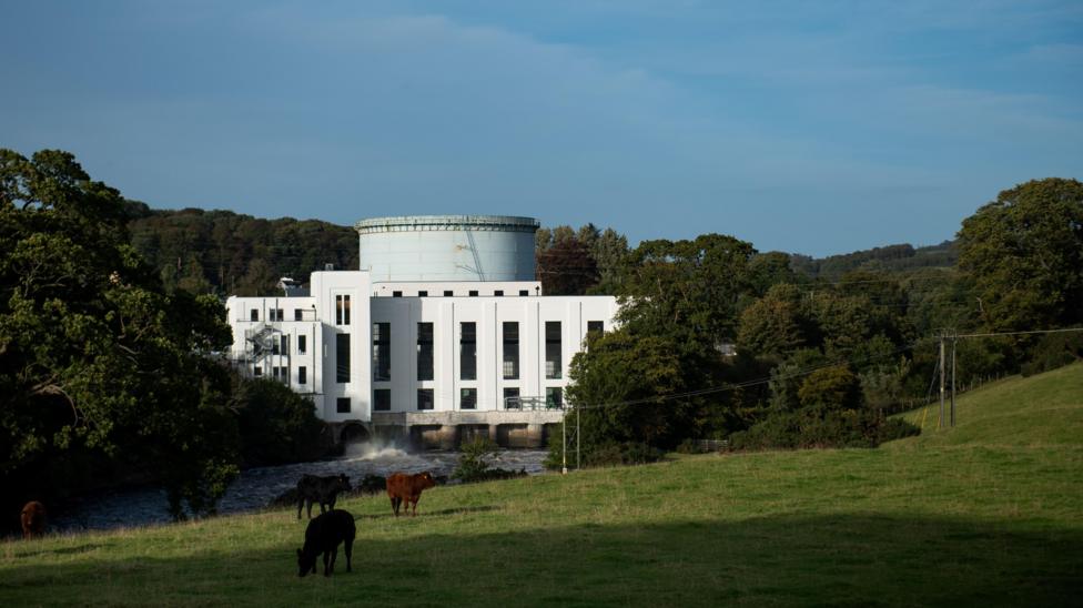 A view of the white art deco Tongland hydro power station building nestling in among trees with cattle grazing in front of it