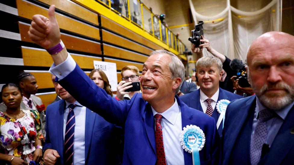 Nigel Farage, smiling broadly, looking up to the left and out of the picture, and holding up an outstretched arm and raised thumb. He is wearing a dark blue jacket, white shirt and patterned red tie. A Reform UK rosette is pinned to his jacket. He is surrounded by other people and photographers.
