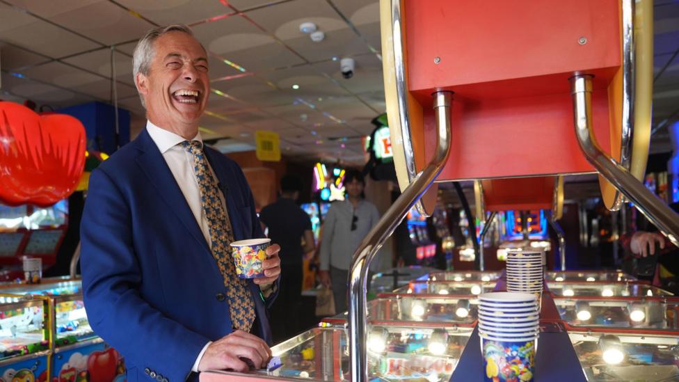 Nigel Farage standing next to a game in an amusement arcade. He is wearing a deep blue jacket, white shirt and patterned tie with what look to be butterflies on it. He is holding a cup, which presumably contains coins. There are other people in the background who are out of focus.