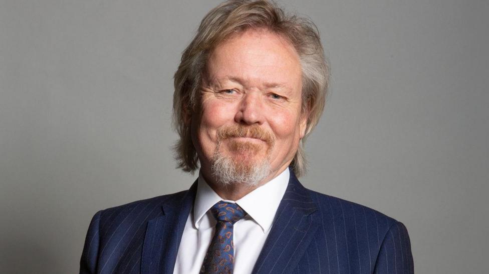 Giles Watling wearing a dark blue, pin-striped suit, white shirt and dark blue patterned tie. He is standing against a plain, grey background. He has a neat goatee beard and light coloured hair that falls over his ears and just reaches his collar.