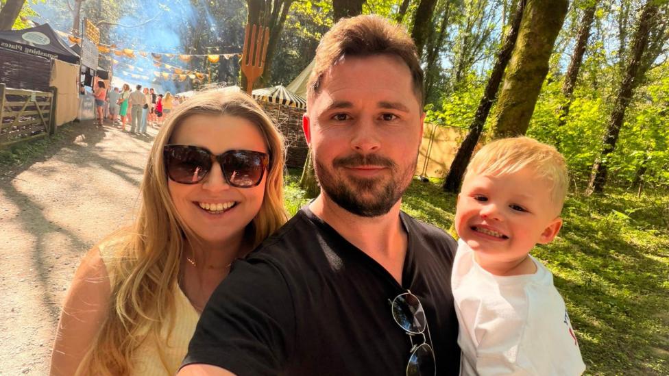 A group selfie from left to right. A woman with large sunglasses and long blonde hair is smiling next to a man with brown beard and hair weaing a black thsir with sunglasses clipped to the collar. He is holding