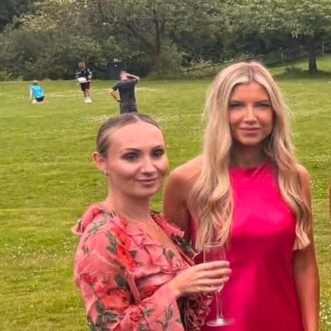 A woman with brown hair tied up is wearing a pink, floral dress and holding a glass of bubbles. She is next to a woman with long blonde hair weaeing a bright pink dress. They are outside, stood on some grass.
