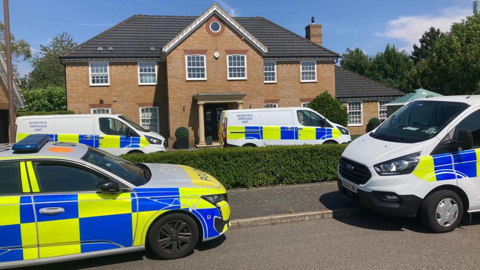 A police car and a few forensic vans are parked outside a large house – some on the street, some on the driveway.
