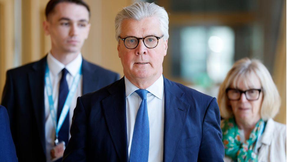 Malcolm Offord, who has grey hair and glasses, looking straight at the camera as he walks down a corridor while wearing a business suit