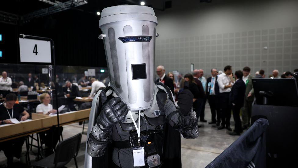 A person in a black costume with a bin for a helmet posing for photos at an election count
