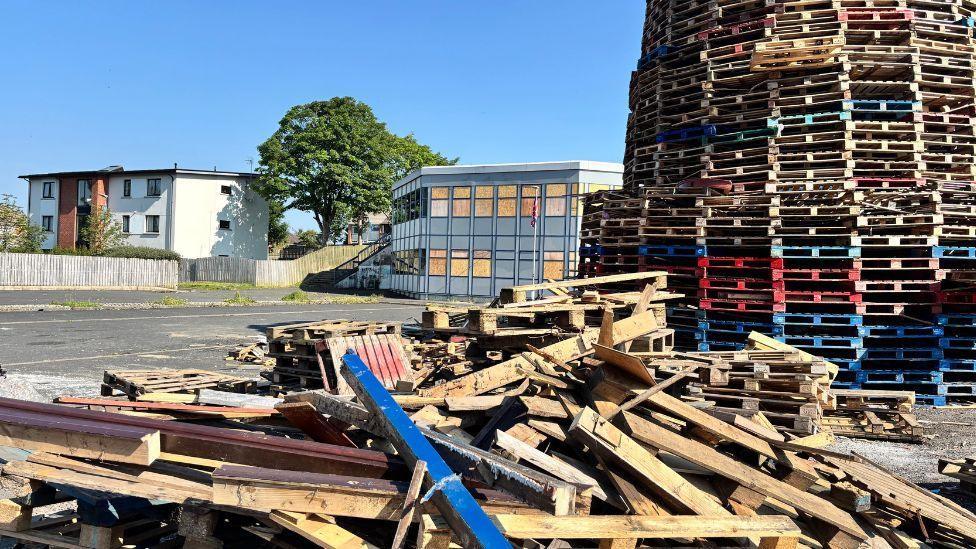 Image of a large bonfire before it is lit - it is constructed from numerous wooden pallets, there is some debris in the front. The library is visible in the background, with boarded up windows