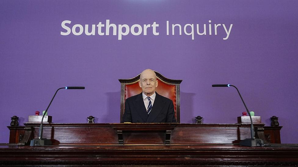 Chair Sir Adrian Fulford sitting inside the hearing room at Liverpool Town Hall, Liverpool, ahead of the start of the southport inquiry. There is a purple background with Southport Inquiry written in white above his desk with two microphones either side of him on an old-fashioned red and wood chair.