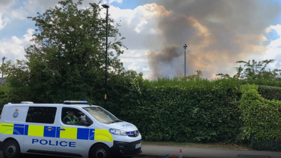 Plumes of smoke behind a hedge. A police car is in front of the hedge.