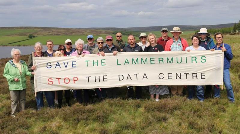 A group of about 20 people standing behind a banner which says Save the Lammermuirs Stop the Data Centre. They are standing in rural countryside with a reservoir behind them.
