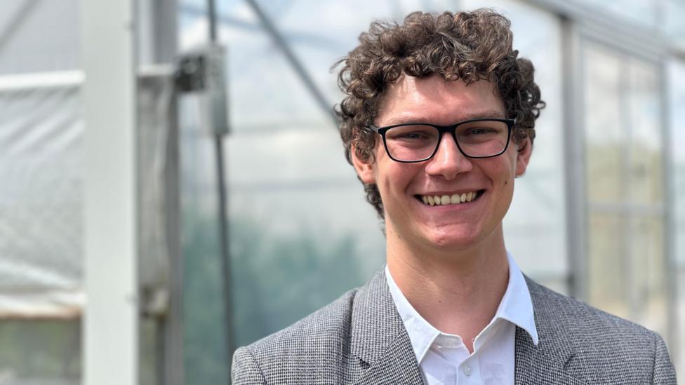 A man wearing glasses, a white shirt with open collar and a grey jacket. He is standing outdoors with what appears to be a greenhouse behind him.