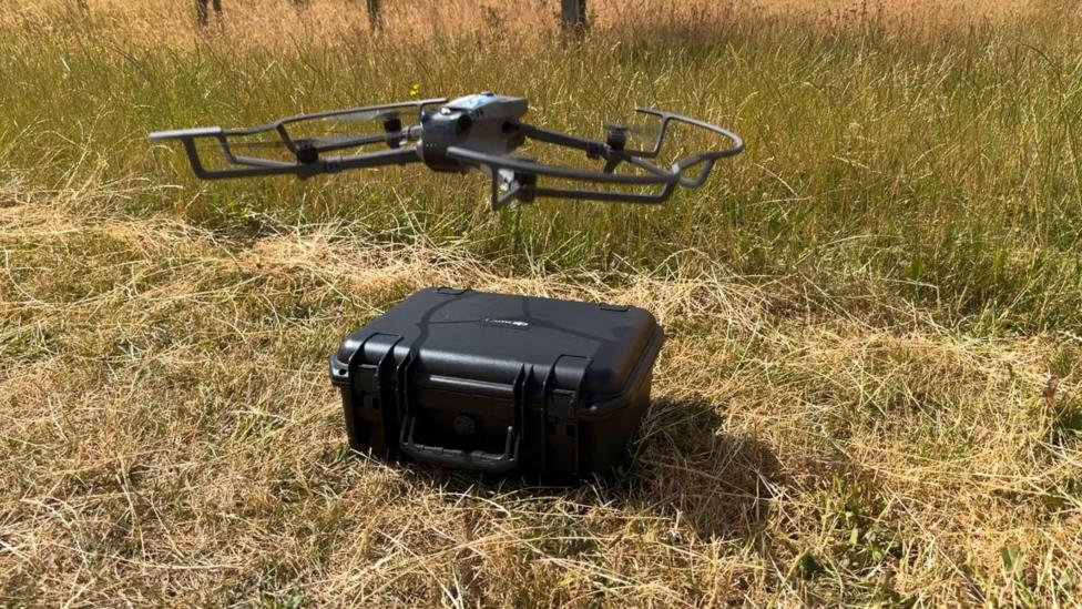 A drone taking off. A black case is on the ground beneath it. They are both located outdoors in an orchard.