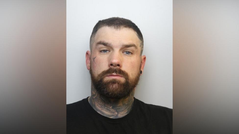 A mugshot of a man in his 30s with dark hair and a beard. He has tattoos all over his neck as well as one on one of his cheeks.