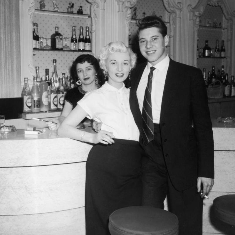 Two women and a man, dressed smartly, standing at a nightclub bar with bottles of alcohol visible in the background. It is a black and white photo and they are all smiling and looking at the camera.