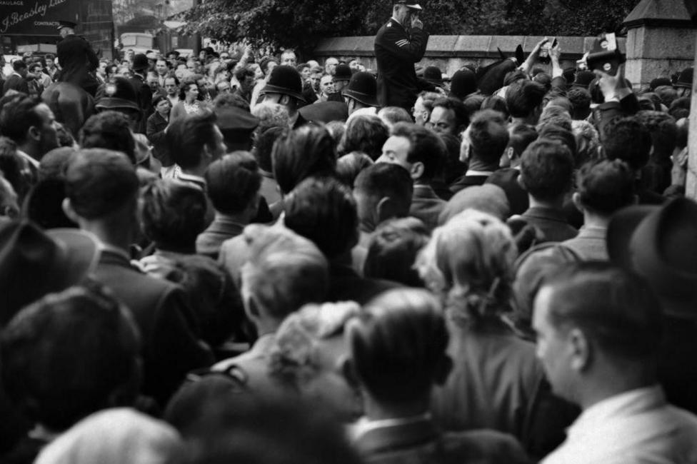 A black and white picture of a tightly-packed crowd of people, from behind. A uniformed police officer is visible stood above them, looking over the crowd.