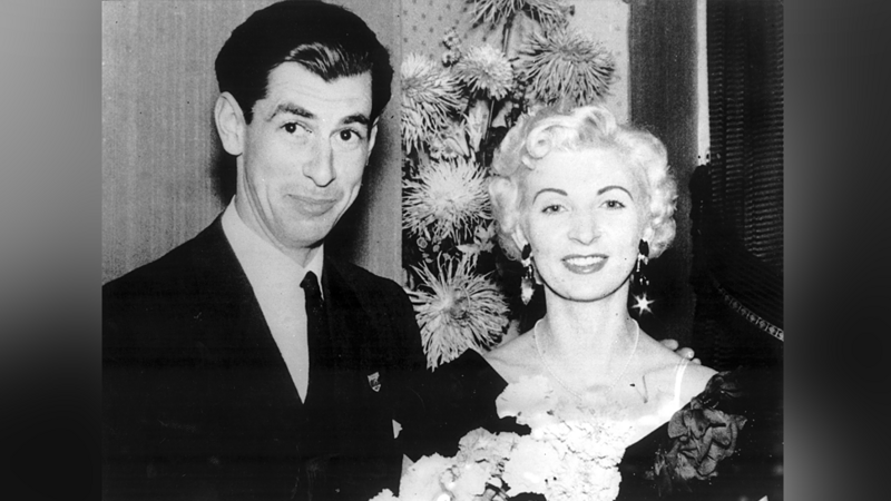 A black and white photo of a tall, dark-haired man and a glamourous blonde woman, with their arms linked. They are both dressed smartly and smile as they look at the camera.