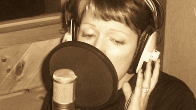 A woman in a studio singing in front of a professional microphone. She has short hair and the picture is in black and white. 