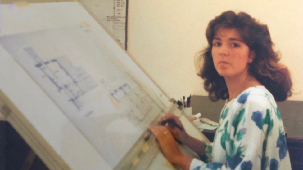A woman with long brown hair sat in front of a board with planning building designs on it. 
