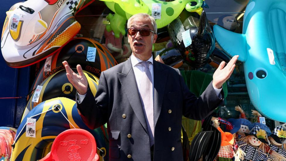Nigel Farage wears a suit and sunglasses. He's standing with his arms stretched to his side, in front of several inflatable toys