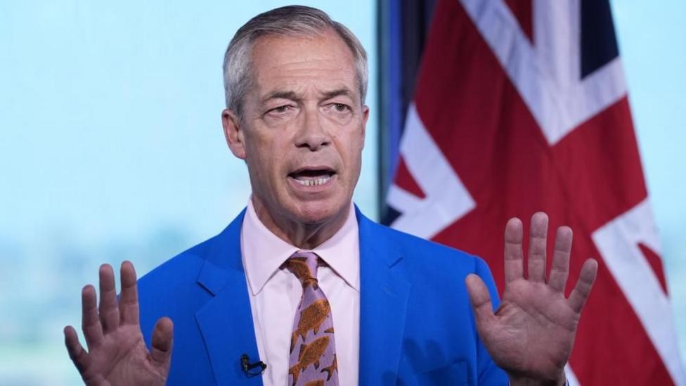 Nigel Farage is wearing a bright blue coloured suit jacket, white shirt and patterned tie. He is stood in front of a Union flag. His hands are held up in front of him.